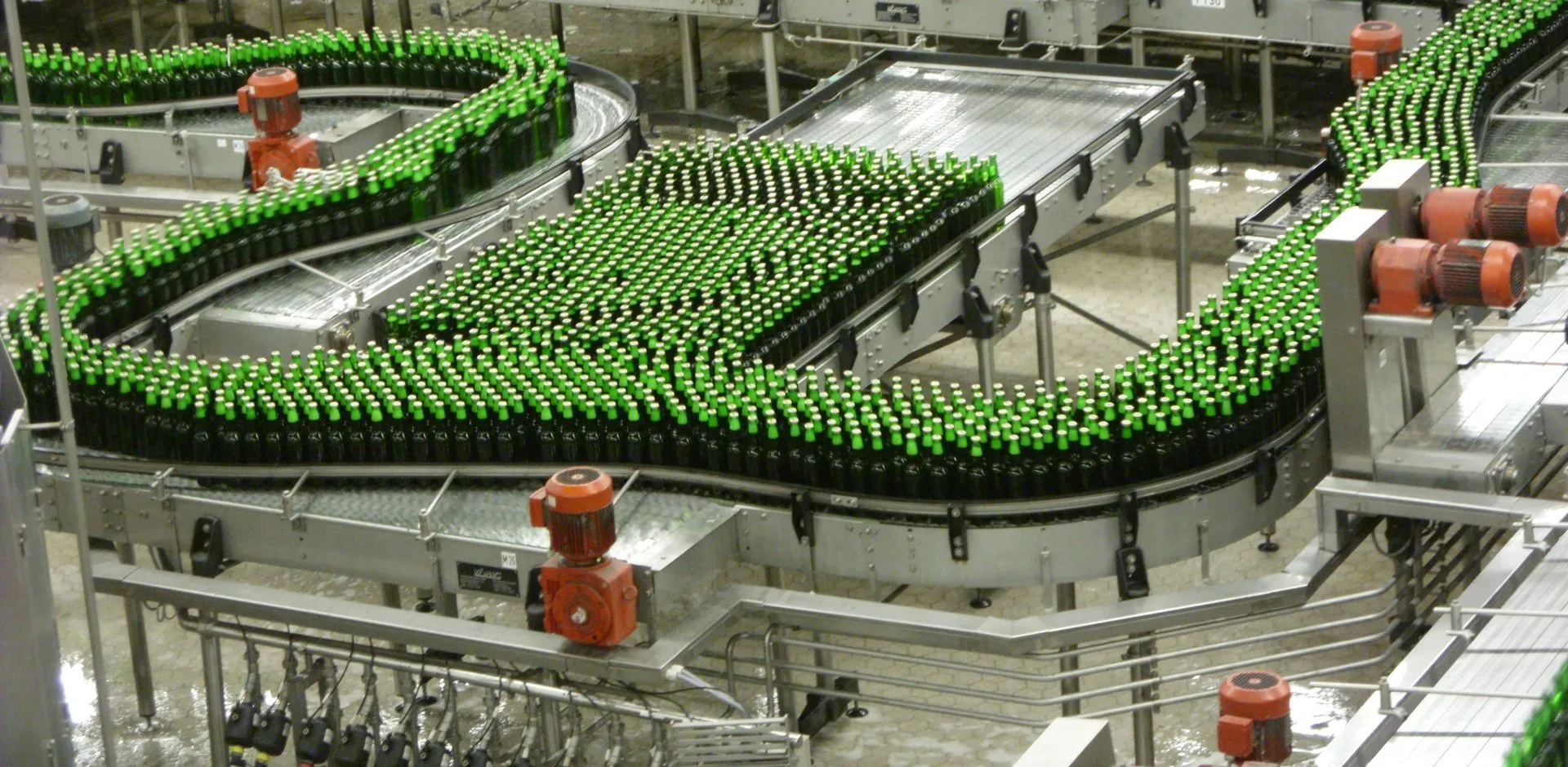 Accumulation conveyors hold product between machines. That buffer is why a short stop on one machine may never reach the filler. Accumulation conveyors carrying thousands of green glass bottles between machines on a brewery bottling line