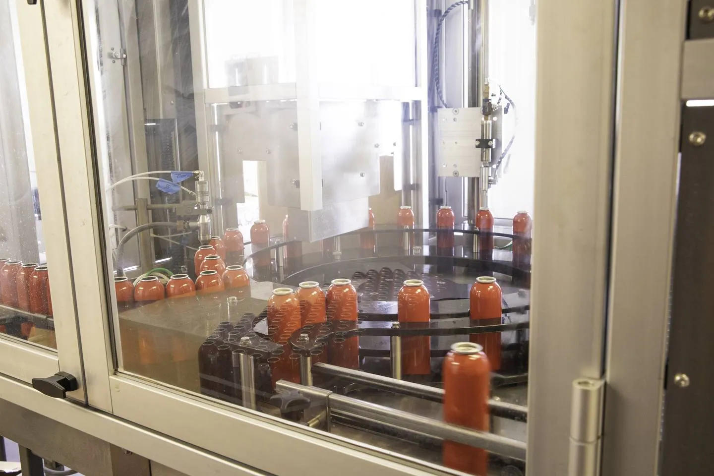Unlabelled red aerosol cans moving around a rotary star-wheel table inside a glass-guarded filling machine