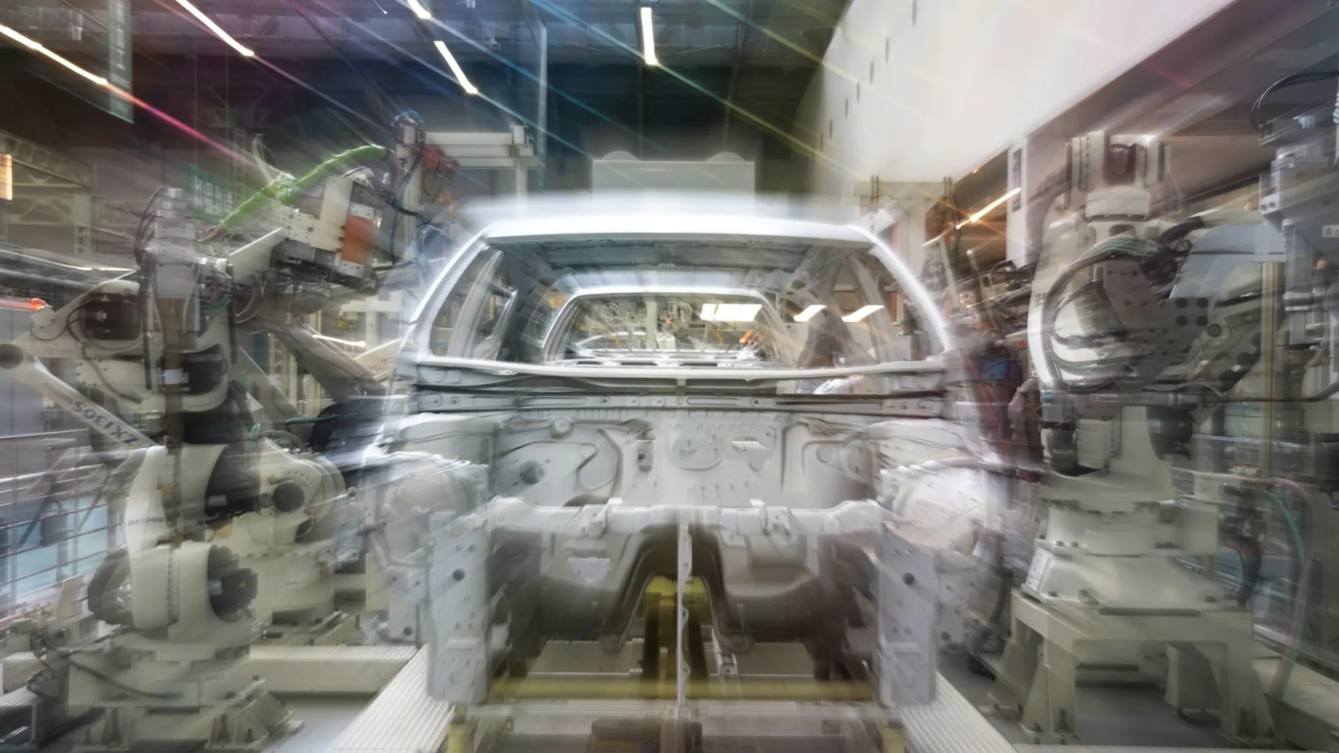 Each robot touch-up in a cell like this is legitimate, and each one ages the last backup. At hundreds of robots per shop, only scheduled uploads keep the backup set worth trusting. Car body shell moving through a robotic welding cell, captured with motion blur