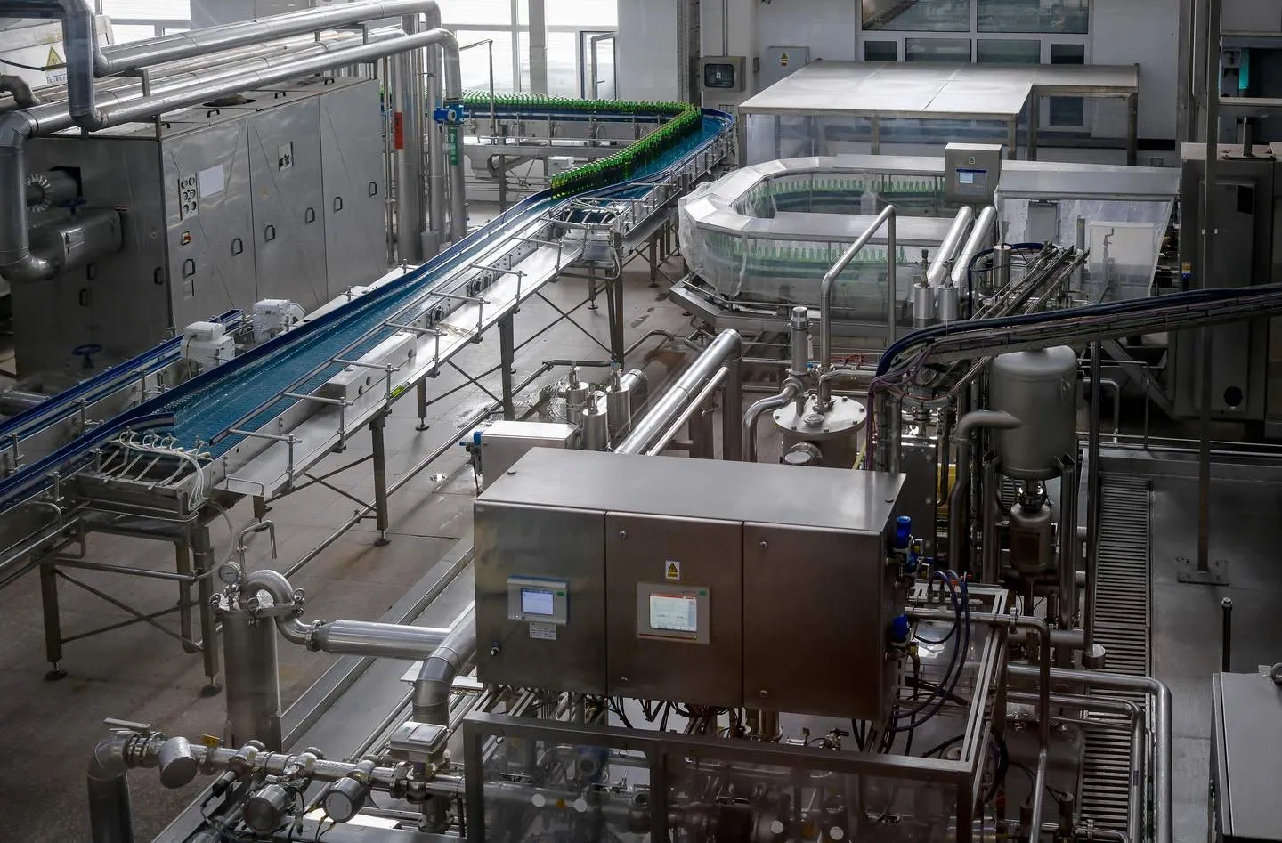 A hybrid plant in one view: a process skid in front, and the bottle conveyors of the packaging half behind it. A beverage bottling hall seen from above, with a stainless process skid and two operator panels in front of conveyors carrying green glass bottles