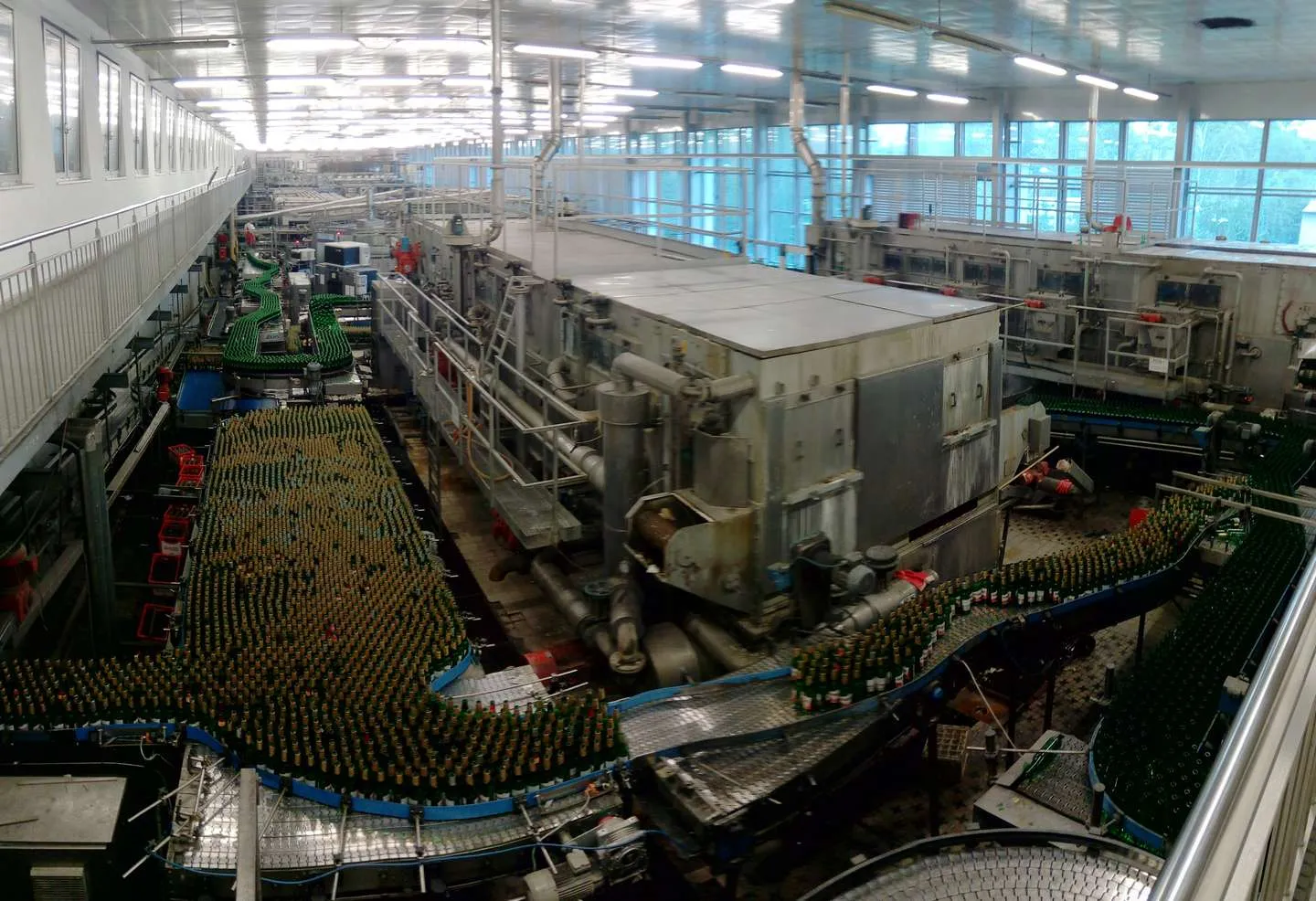 A brewery bottling hall. Bottles queue on the conveyors before and after the tunnel pasteurizer in the middle, the accumulation that keeps a short stop at one machine from stopping the line. A brewery bottling hall seen from a gallery, with thousands of green bottles on curved conveyors around a large stainless tunnel pasteurizer