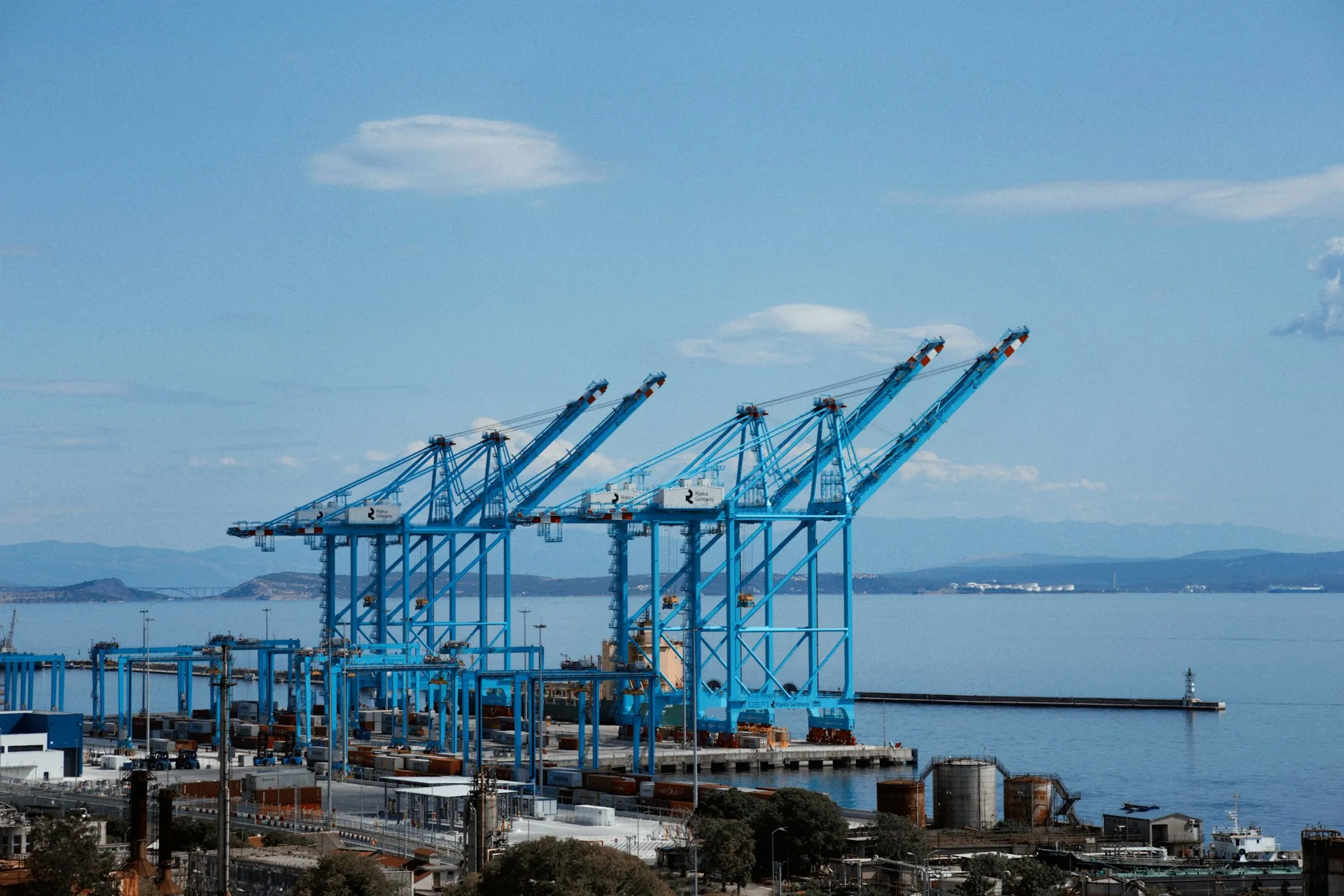 A port moves cargo because every crane, truck and ship hands off through a shared standard. Micro-integrations play that role between business systems and the event broker, so no system has to know about the others. Row of blue ship-to-shore gantry cranes at a container port under a clear blue sky