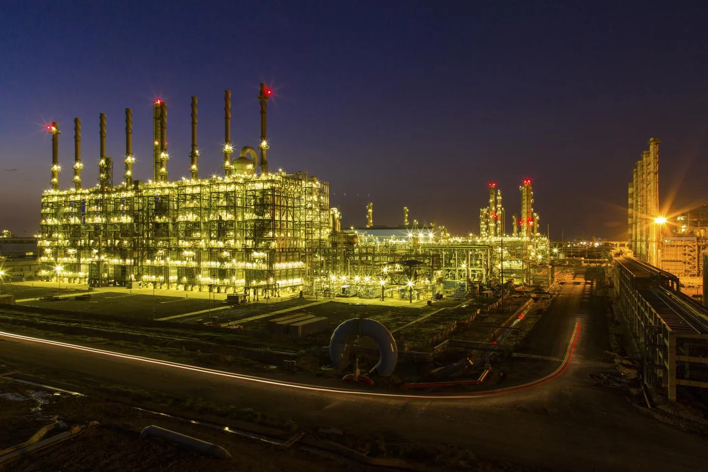 A cracker furnace block with a row of tall stacks, lit up at blue hour, with pipe racks and a road in the foreground