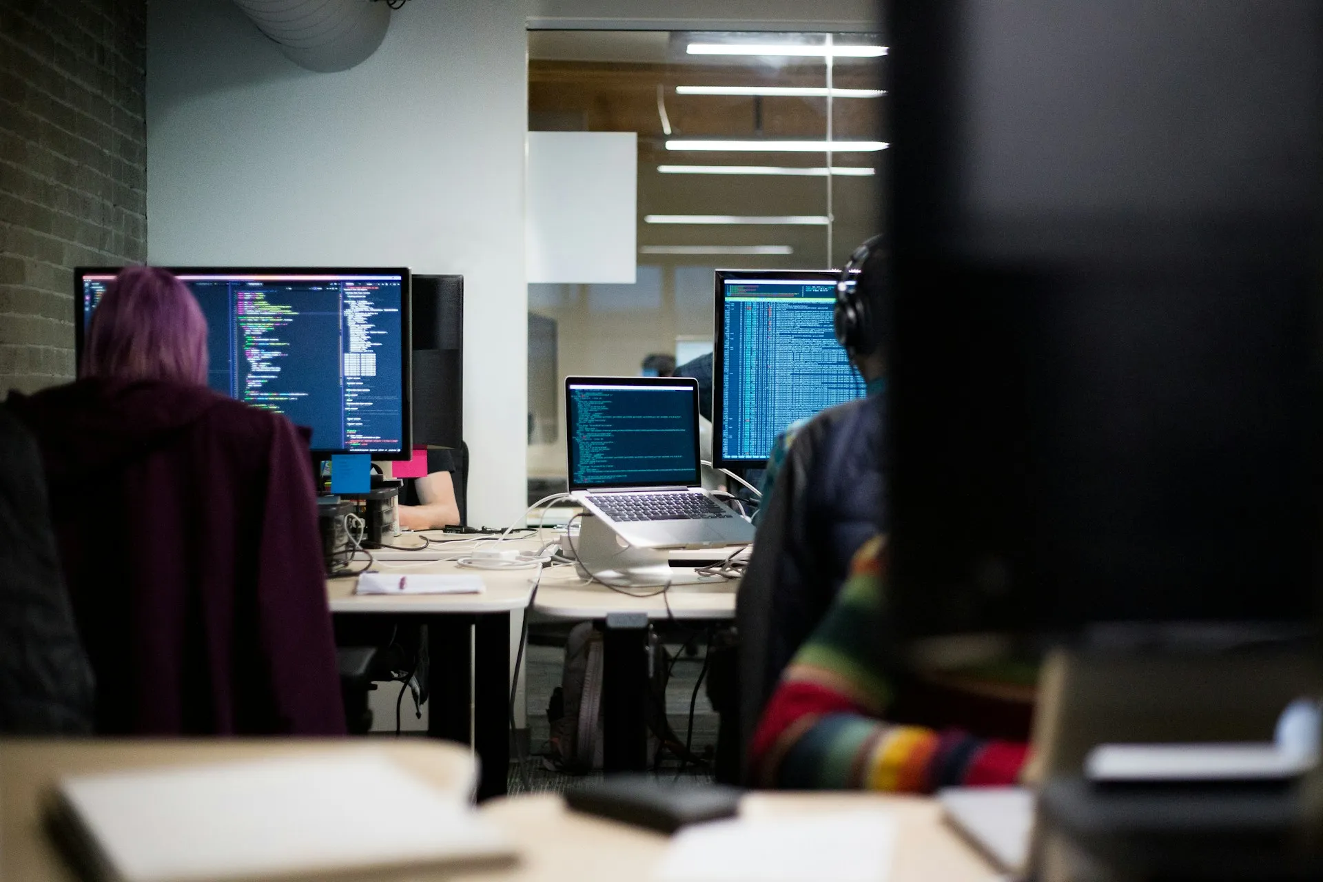 Copia brings a software team's habits to PLC code: commits, branches, reviews. That pays off where engineers already work this way, and adds a skill to learn where they do not. Developers at desks with code-filled monitors and a laptop in an open-plan office