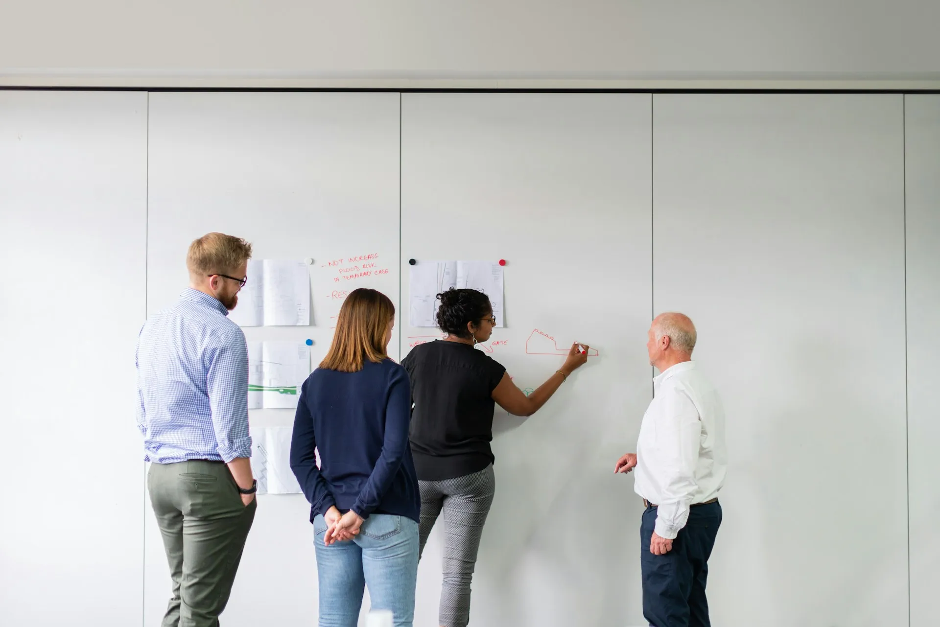 Agree the cutover order before any software moves: which area goes first, what the pilot must prove, and who signs off each batch. Four engineers at a whiteboard wall with pinned drawings while one sketches the next step in red marker