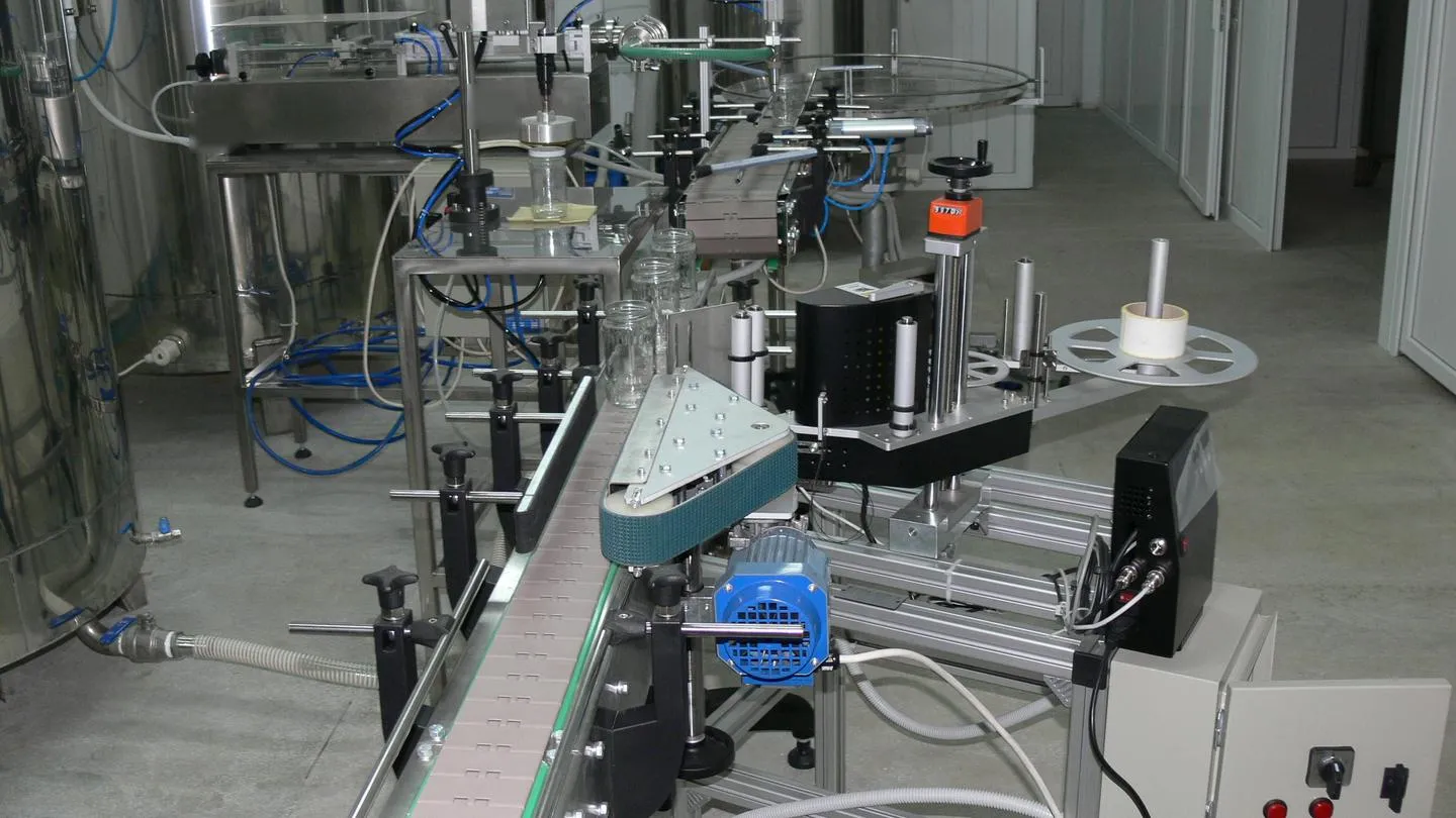 A small filling, capping and labelling line, with adjustable conveyor guide rails on star-knob clamps, a labeller with a hand wheel and counter, and stainless tanks behind