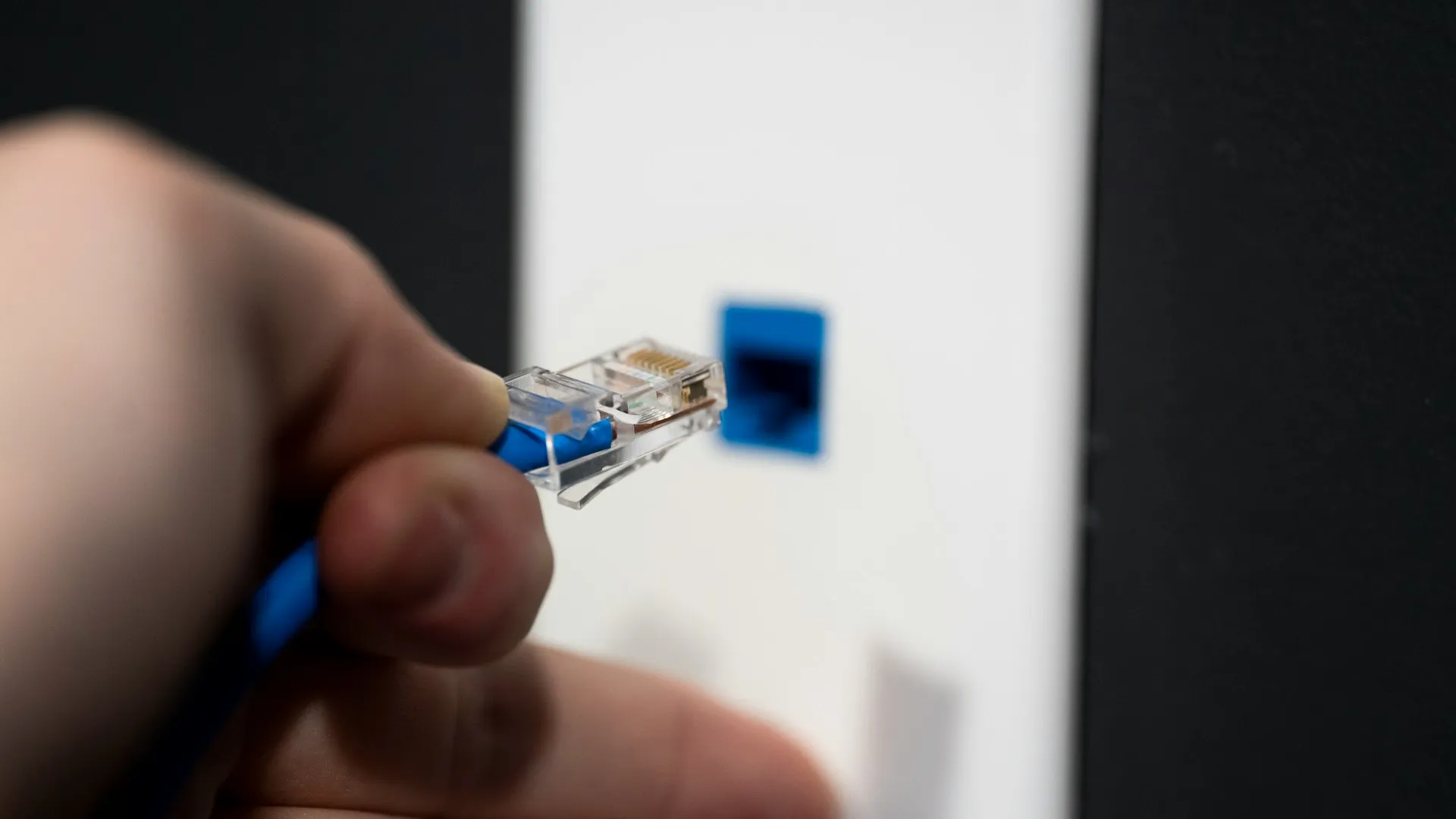 The plug and the cable are ordinary Ethernet. What makes it EtherNet/IP is the CIP traffic running over them, and that traffic does not tolerate what office traffic shrugs off. Hand holding a blue patch cable's RJ45 plug in front of a blue network wall port
