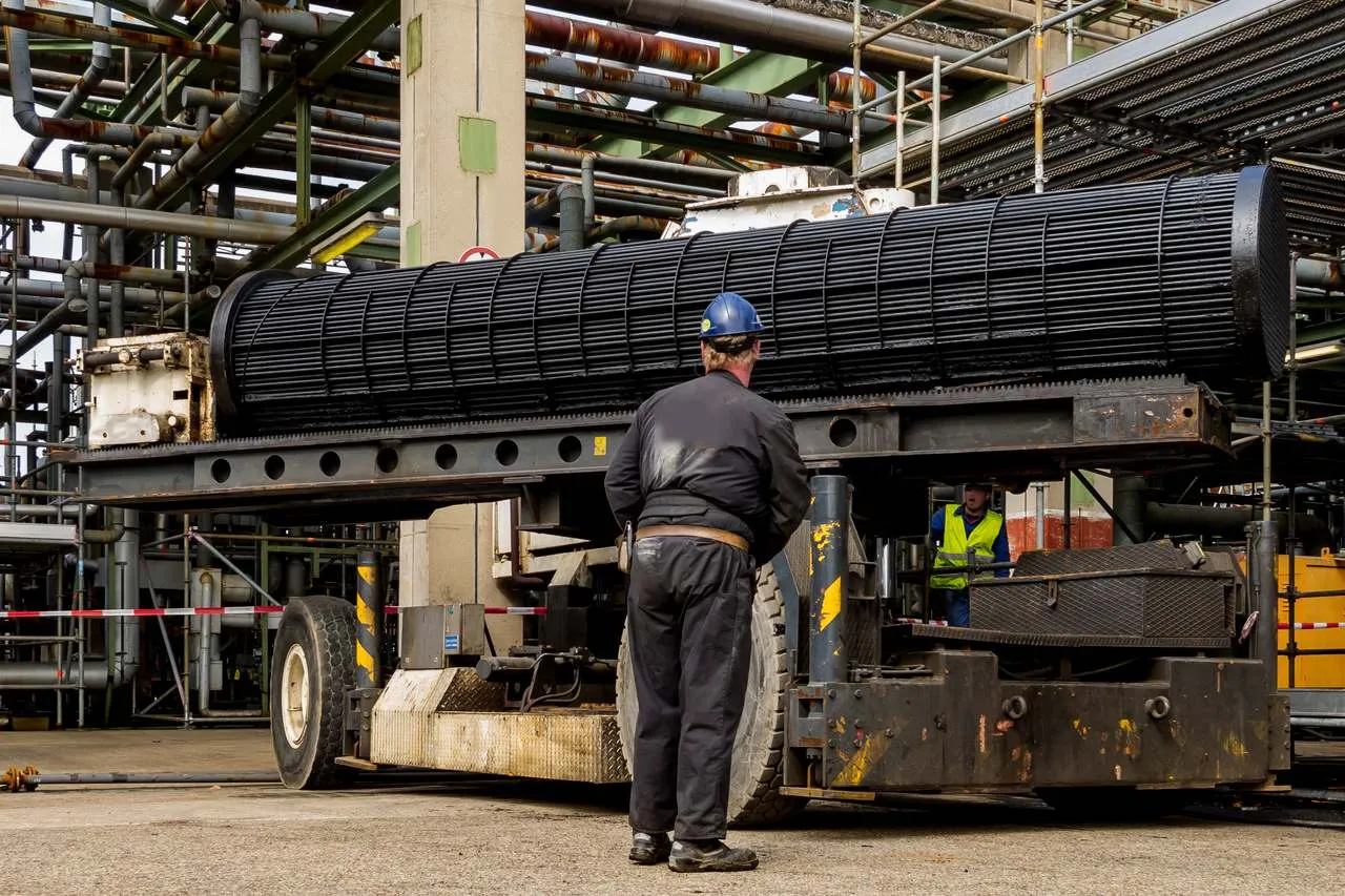 A heat exchanger tube bundle, pulled out of its shell, resting on an extractor carrier between pipe racks, with a worker in a hard hat standing beside it