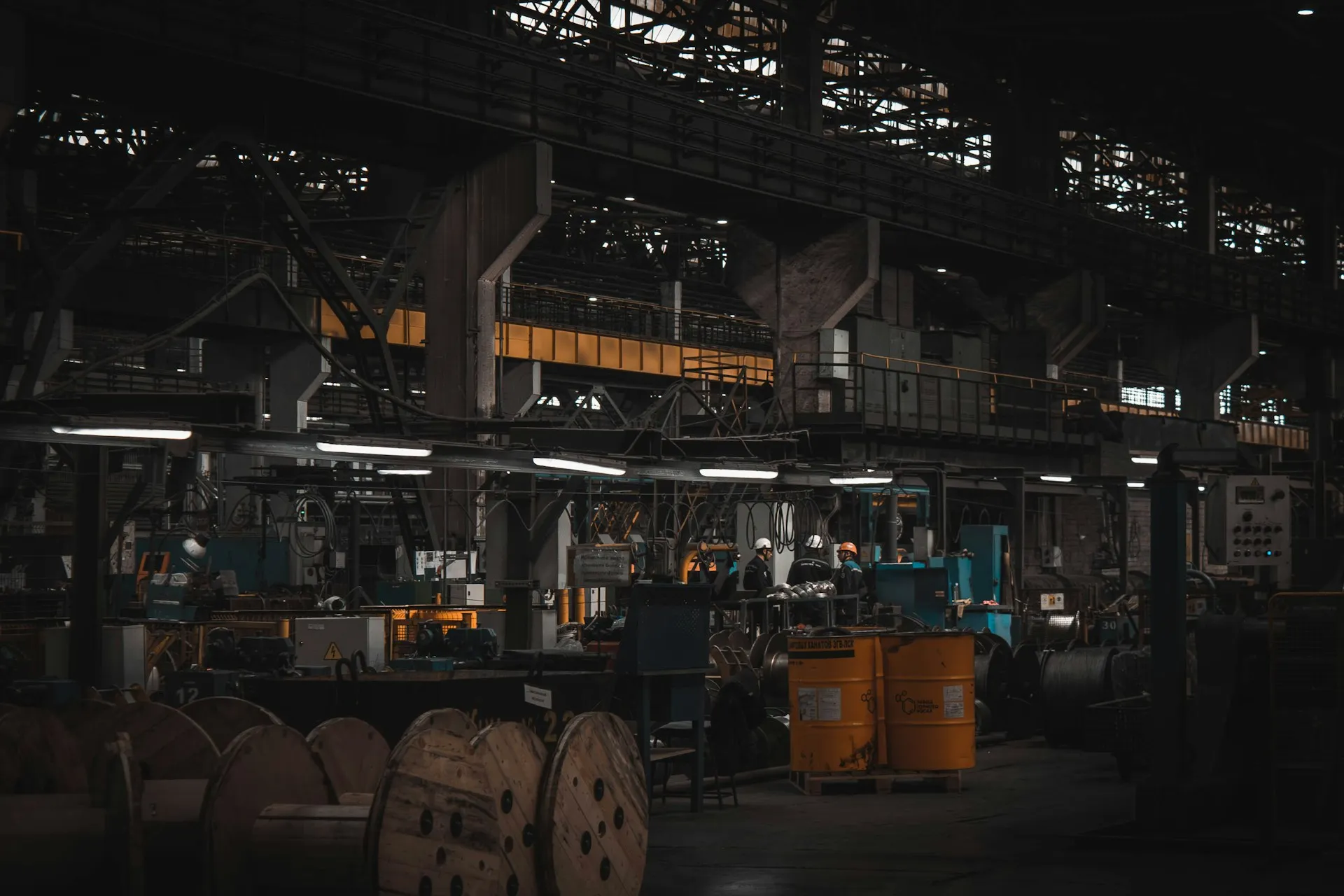 On a floor like this, recovery time depends on one thing nobody can see: whether a current, trusted copy of every program exists. That is the record AMDT's products are built to keep. Dim heavy industrial plant floor with cable reels, oil drums and machinery under steel roof trusses