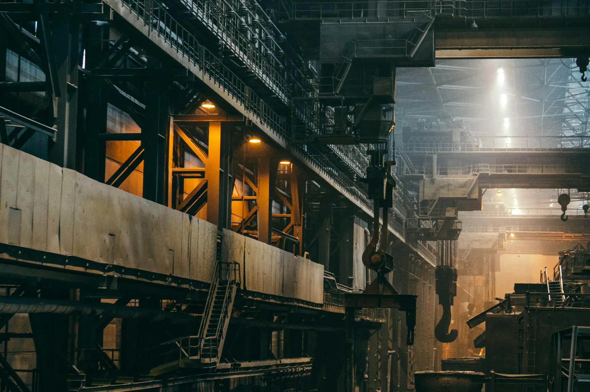 Large heavy-industry plant hall with steel structure, overhead cranes and hooks in hazy light