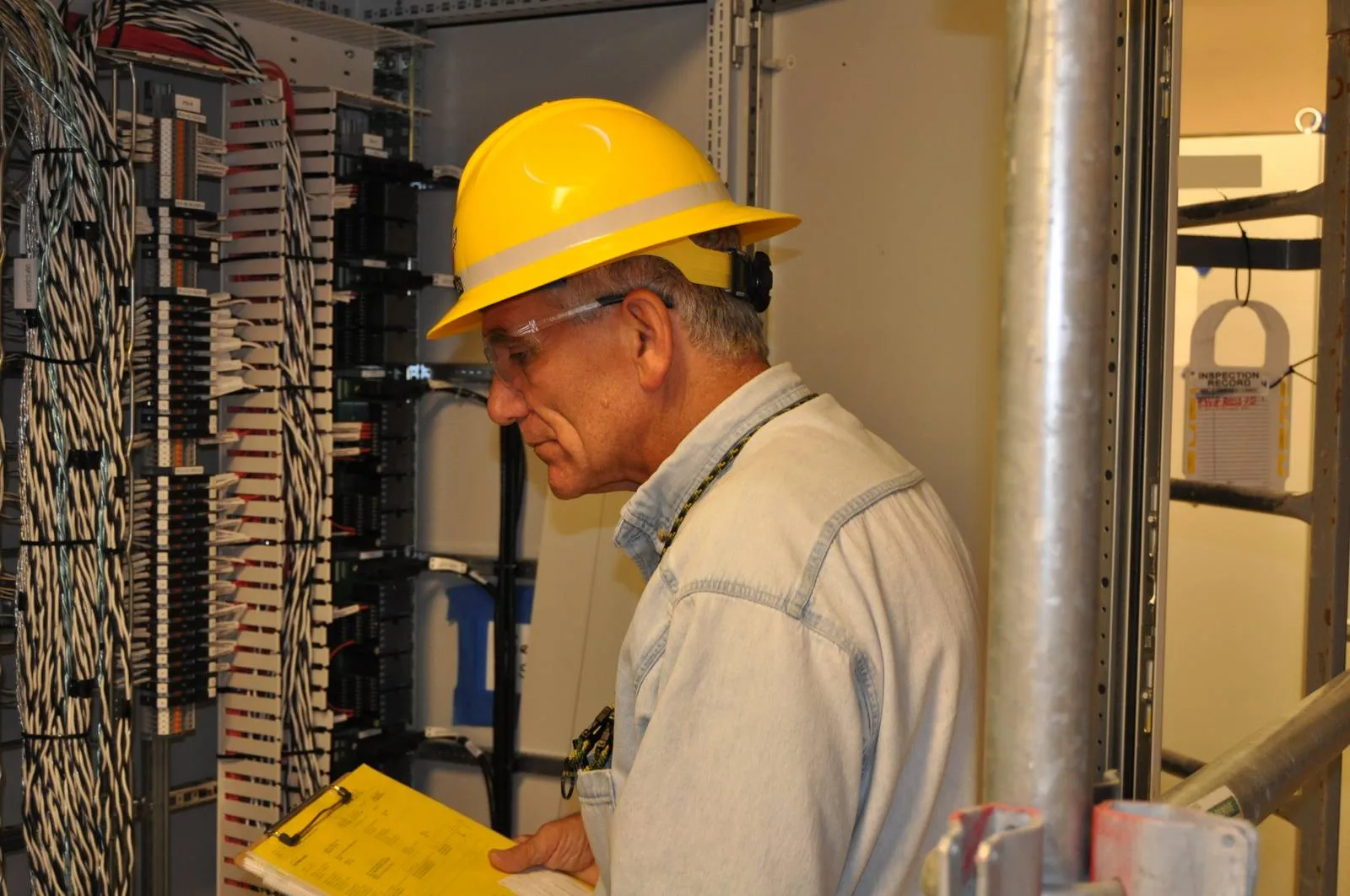 Security acceptance tests belong on the same checklist as every other test, run before the system is handed over. Inspector in a yellow hard hat checks wiring in a control cabinet while holding a clipboard checklist