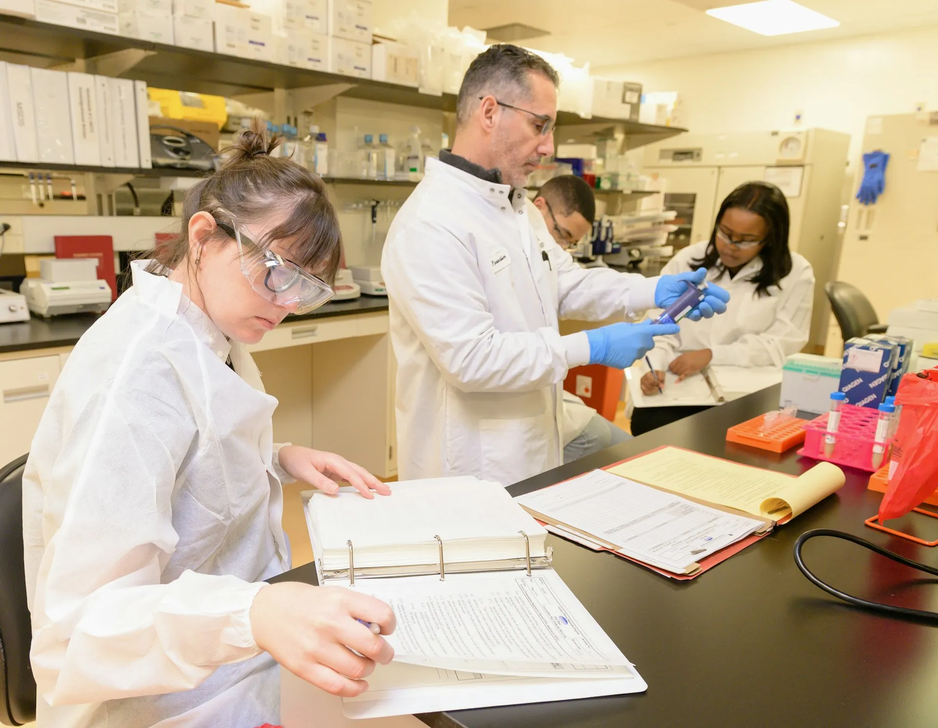 An auditor samples both kinds of record: the tool supplies the dated versions and change history, while the procedures, approvals and training files still come from the plant's own quality system. Laboratory technicians in white coats reviewing a records binder and handling sample tubes at a bench