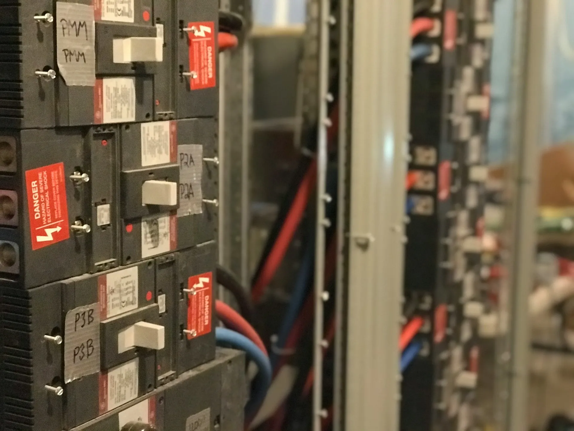 Panels built by different integrators over different decades are the norm. The more of them run on non-Rockwell controllers, robots and drives, the more a multi-vendor tool earns its place. Hand-labelled circuit breakers with danger stickers inside a row of industrial control panels