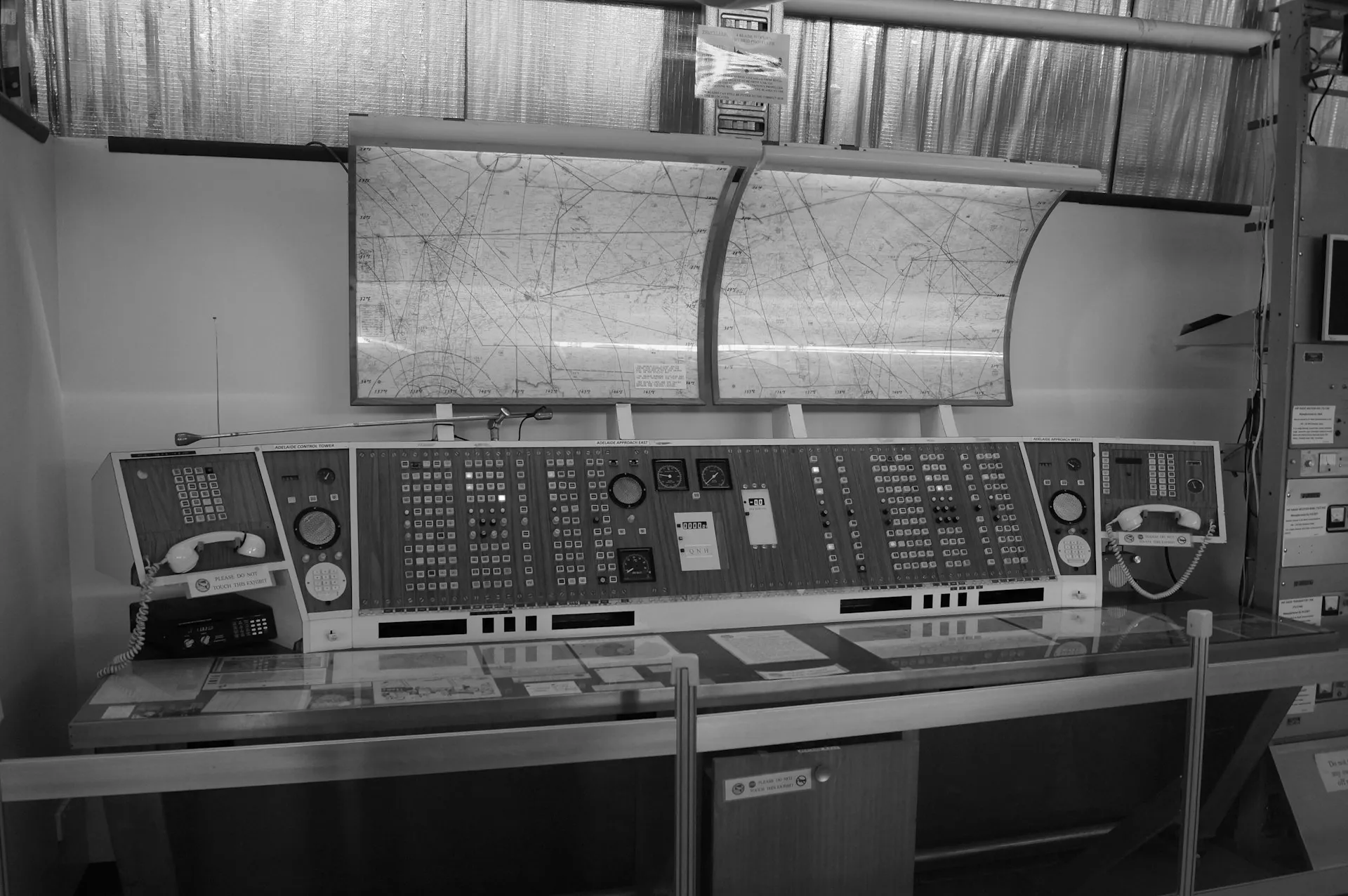 Multi-site visibility is the old mimic-board idea applied to asset data: one board for every plant, so a question about exposure is answered by looking, not by emailing eight sites. Black and white control room desk with switch panels and telephones beneath a large mimic map