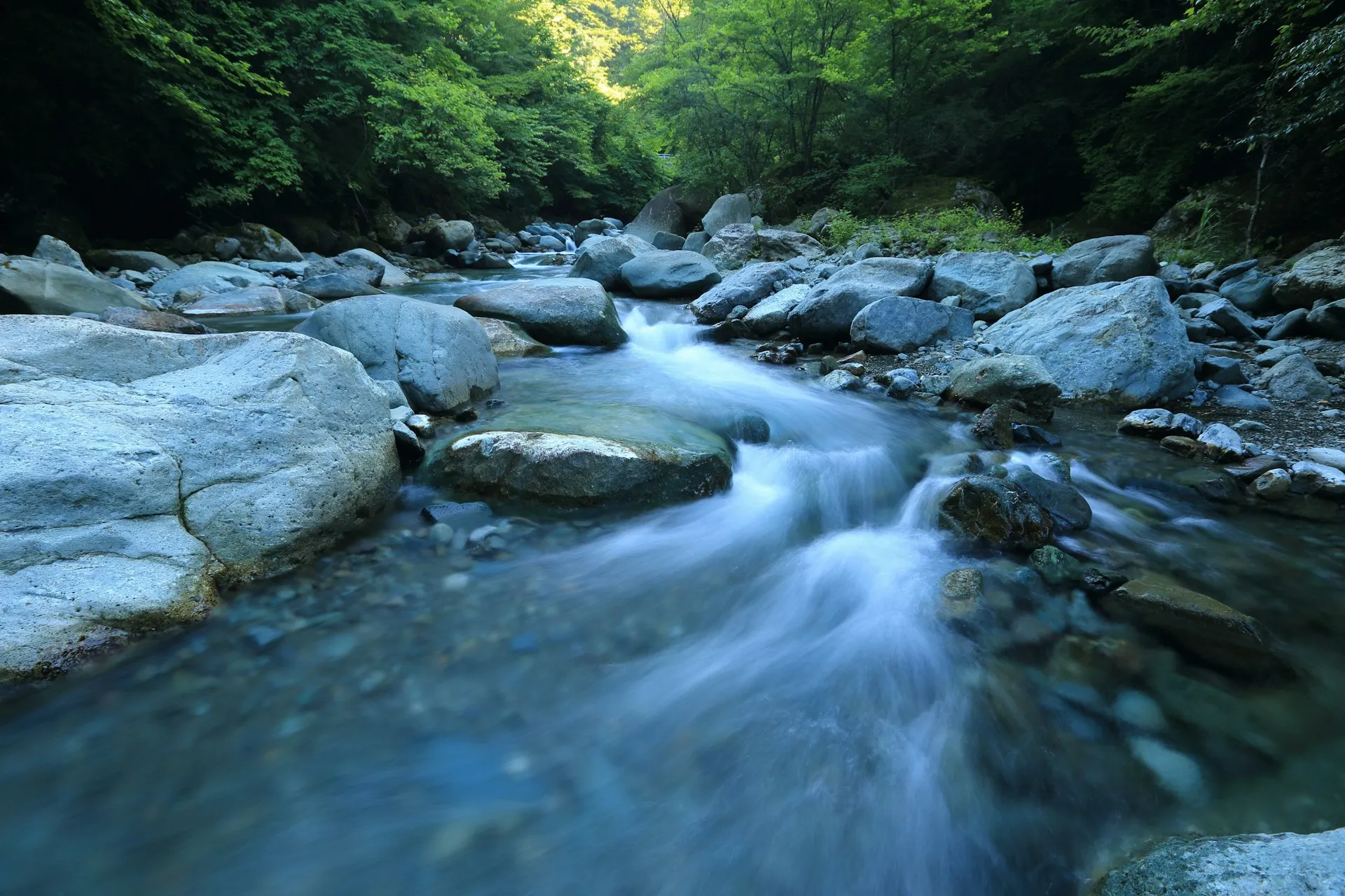 A stream keeps flowing whether or not anyone is drawing from it, which is Kafka's model; a broker instead directs each event to the consumer that asked for it. Long exposure of a clear mountain stream rushing over smooth grey boulders in a green forest