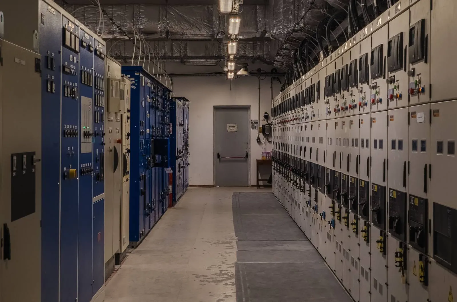 Most plants run several generations of equipment side by side. A 62443 program has to protect all of them, not only the newest. Long aisle of older blue and newer beige switchgear and control cabinets in a plant electrical room