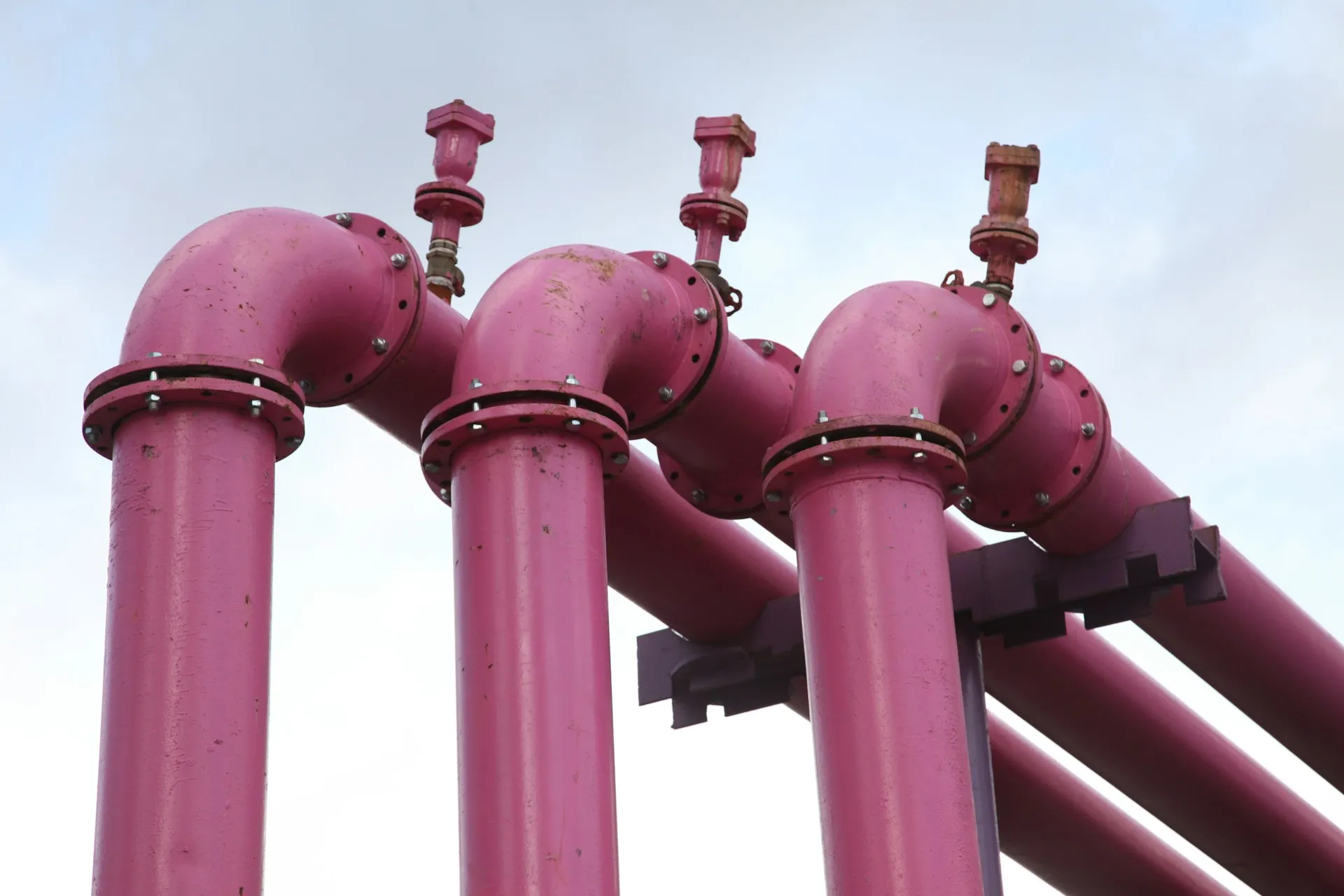 Each connector is a junction where one system's flow joins the shared network. Its configuration decides what passes through, in what shape, and what happens when the downstream side stops. Three magenta painted pipes with elbow joints and valves running in parallel against a pale sky