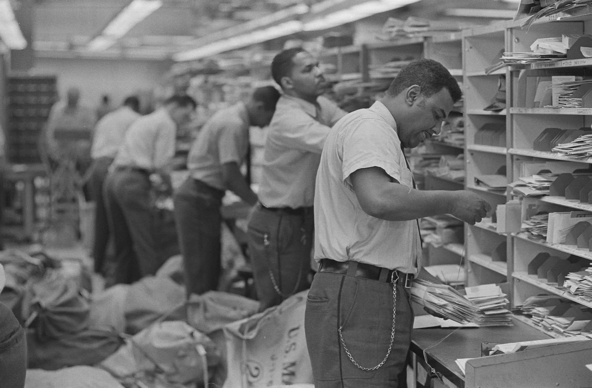 Senders write an address, and the sorting room decides who gets a copy. Topics are the address; subscriptions and queues are the pigeonholes. Black and white photo of postal workers sorting letters into rows of pigeonhole shelves