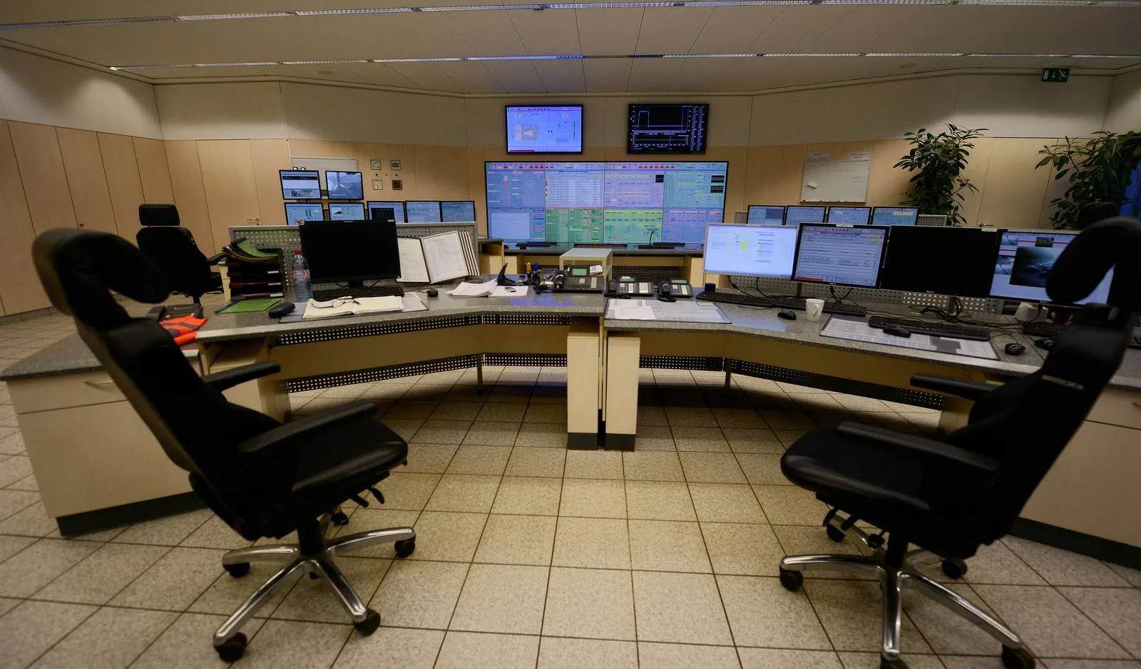 A coal-fired power station control room in Germany. In process industries, operators run the plant from consoles like these. Unoccupied operator consoles in a power station control room, with a wall of process display screens behind them and two chairs in the foreground