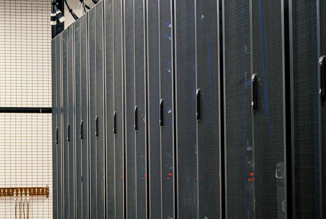 Server racks. The AssetCentre server and its SQL Server database hold every archived program, so where they run, and who can reach them, matters. A row of black mesh-door server cabinets beside a wire-mesh cage wall in a server room