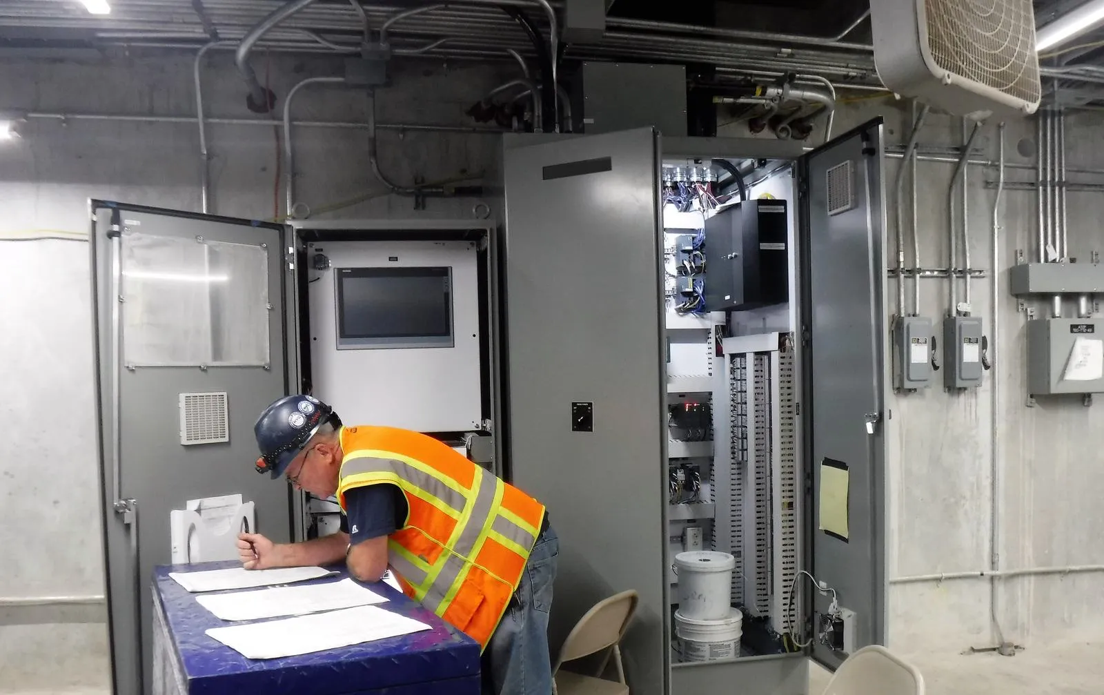 Technician reviewing printed checklists on a toolbox beside open control cabinets with an HMI and PLC wiring