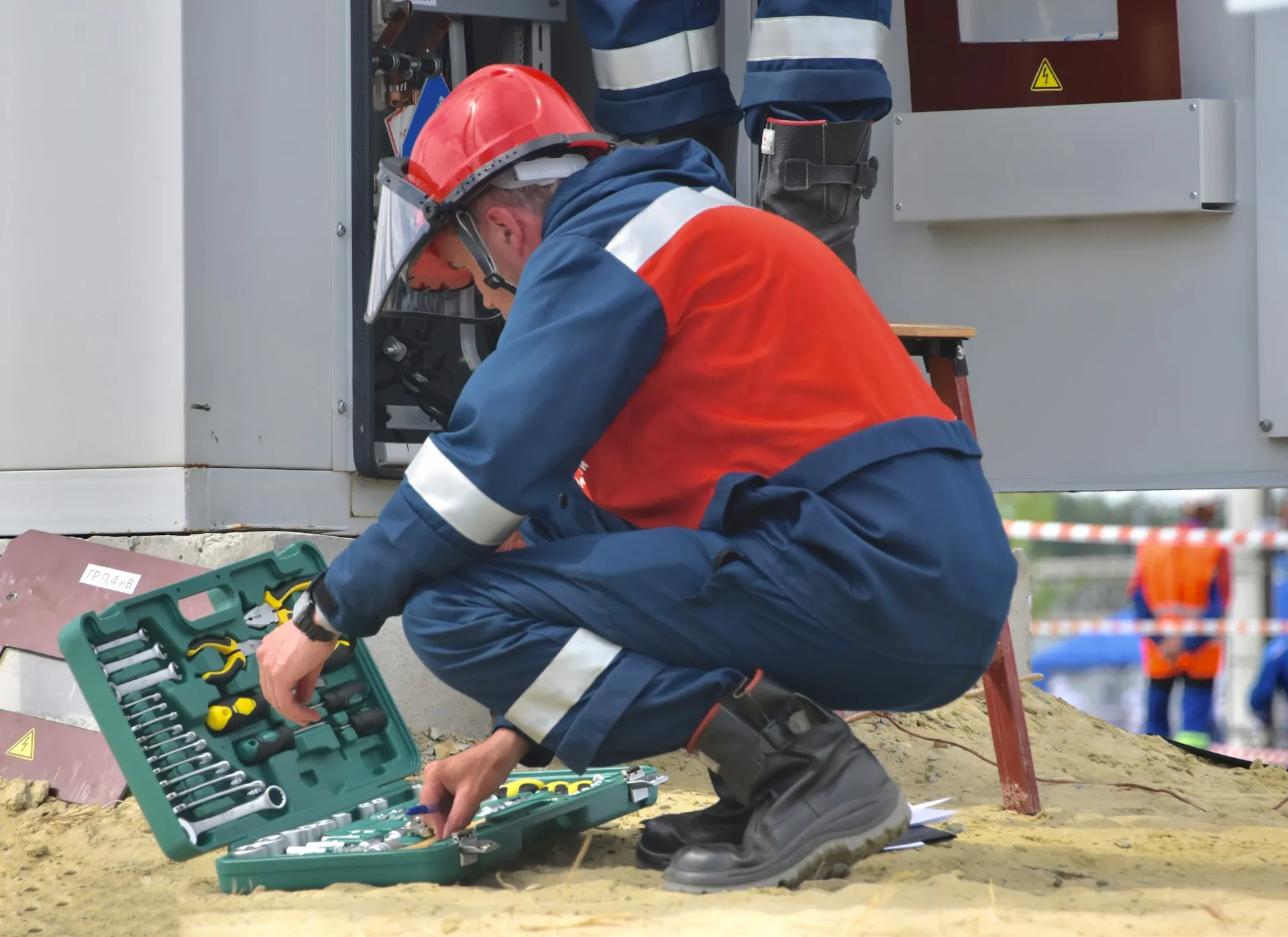 A replacement drive or controller fitted today is protected only if the backup tool can read its firmware and project format, and versiondog stopped learning new formats in 2025. Technician in red hard hat and face shield kneeling at an open electrical cabinet with an open toolkit