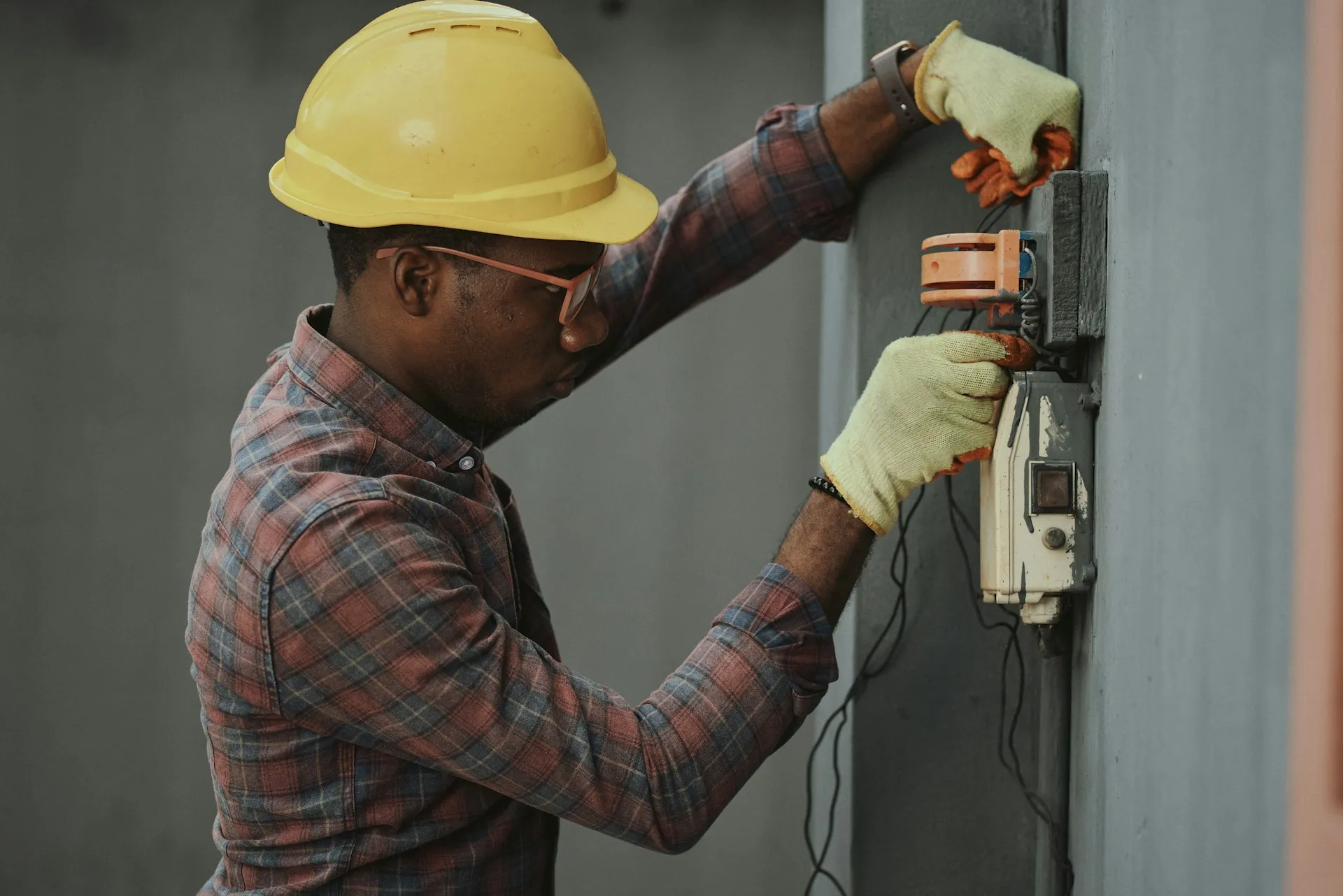 Scans and backups produce a list; someone standing at the panel is what confirms the list matches the plant. Technician in hard hat and gloves inspecting the wiring of a wall-mounted electrical device