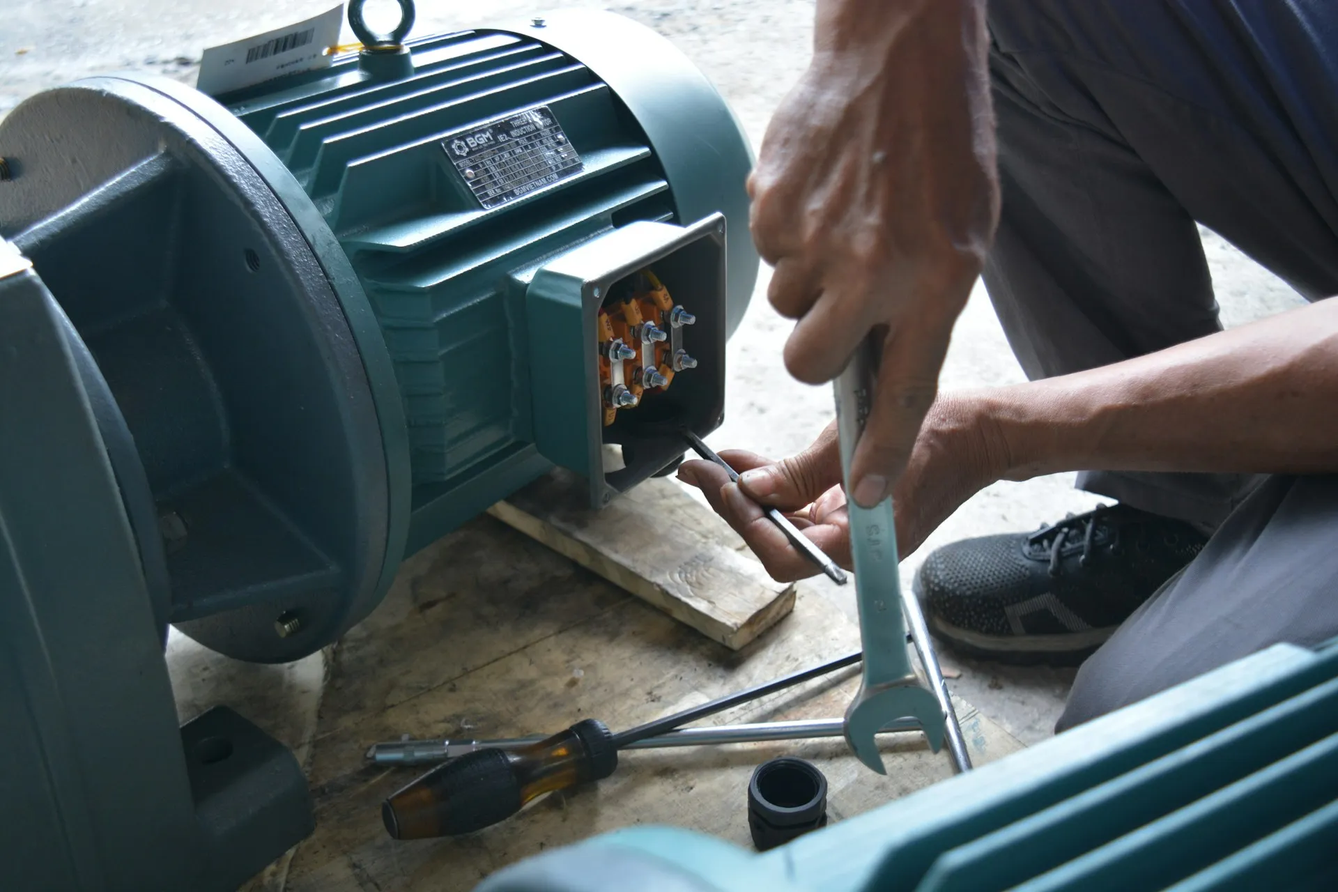 OT changes are made with hand tools and laptops at the machine, often on a night shift, so the change record has to be captured from the device rather than typed into a ticket afterwards. Technician using a wrench and screwdriver on the terminal box of a large industrial motor