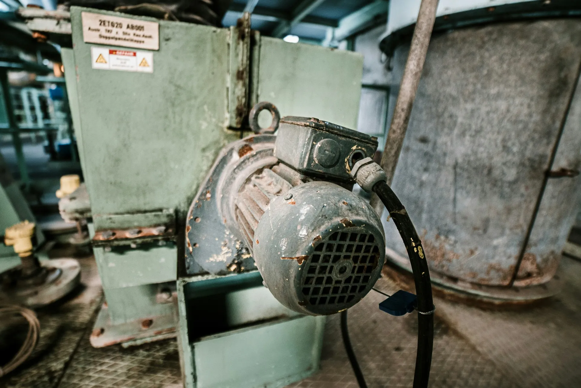 Many trips logged against the drive start at the motor, the cable or the load it is turning. Look at the motor and its cable gland before ordering a replacement drive. Weathered induction motor with its supply cable bolted to a green hopper in an old processing plant